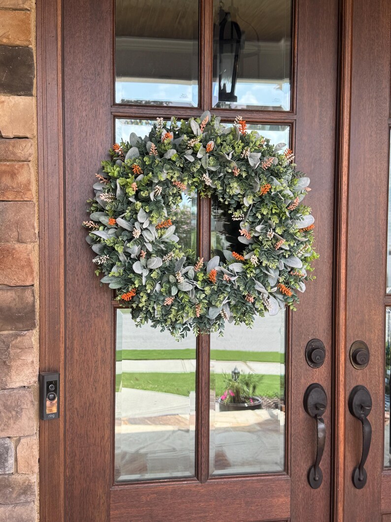 May include: A circular wreath featuring a dense arrangement of green eucalyptus leaves, soft grey lamb's ear foliage, and small clusters of bright orange floral accents, mounted on a dark brown wooden door with glass panels and metal hardware.