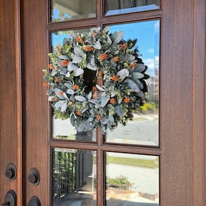 May include: A circular wreath featuring soft, sage green lamb's ear leaves interspersed with small, vibrant orange floral accents. The wreath is mounted on the glass pane of a dark brown wooden door with visible metal door handles and hardware.