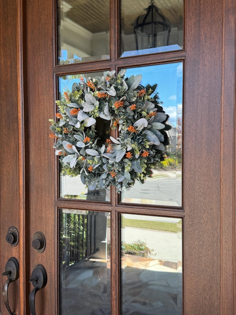 May include: A circular wreath featuring soft, sage green lamb's ear leaves interspersed with small, vibrant orange floral accents. The wreath is mounted on the glass pane of a dark brown wooden door with visible metal door handles and hardware.