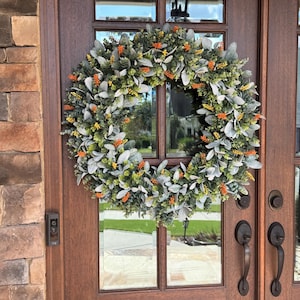 May include: A circular wreath composed of silver-green lamb's ear leaves, small green foliage, and scattered bright orange and yellow floral accents. The wreath is mounted on a dark brown wooden door with glass panels and metal hardware.