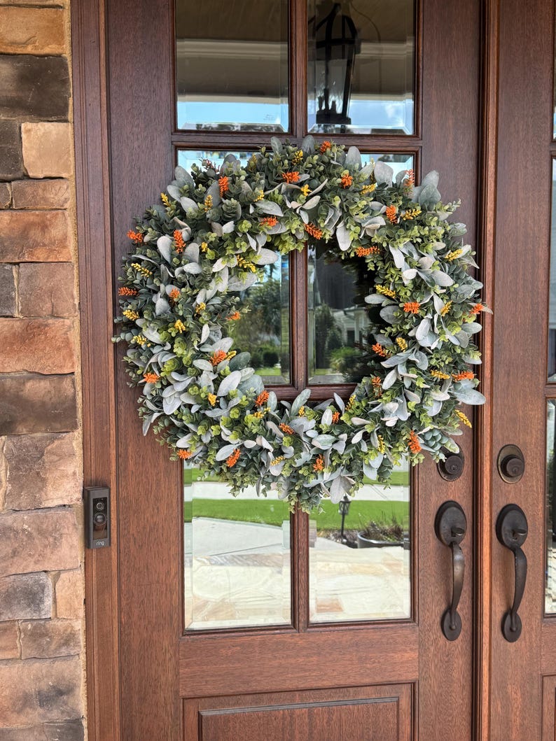 May include: A circular wreath composed of silver-green lamb's ear leaves, small green foliage, and scattered bright orange and yellow floral accents. The wreath is mounted on a dark brown wooden door with glass panels and metal hardware.