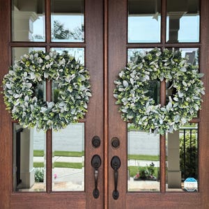 May include: Two circular wreaths made of green foliage and silver-toned lamb's ear leaves are mounted on a set of dark brown wooden double doors with glass panels. Each door features a black metal handle and a matching deadbolt lock mechanism.