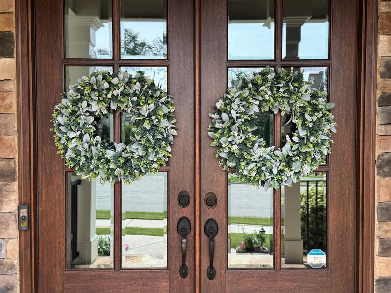 May include: Two circular wreaths made of green foliage and silver-toned lamb's ear leaves are mounted on a set of dark brown wooden double doors with glass panels. Each door features a black metal handle and a matching deadbolt lock mechanism.