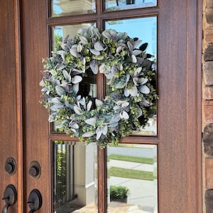 May include: A circular wreath featuring a dense arrangement of sage green lamb's ear leaves, small green eucalyptus sprigs, and clusters of purple lavender flowers. The wreath is mounted on the glass panel of a dark brown wooden door with metal hardware.