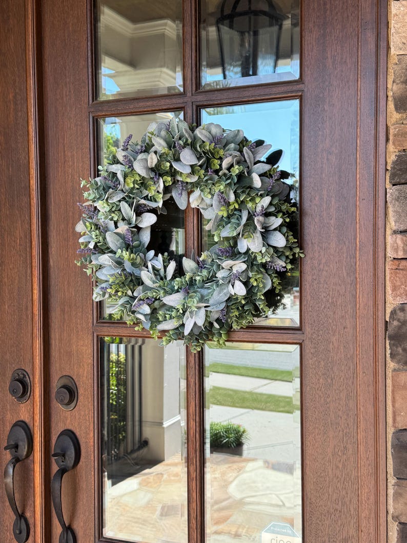May include: A circular wreath featuring a dense arrangement of sage green lamb's ear leaves, small green eucalyptus sprigs, and clusters of purple lavender flowers. The wreath is mounted on the glass panel of a dark brown wooden door with metal hardware.
