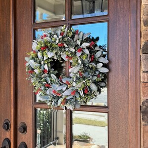 May include: A circular wreath featuring a dense arrangement of soft, sage-green lamb's ear leaves interspersed with small, vibrant red berry clusters and sprigs of green foliage, mounted on a dark brown wooden door with glass panels.