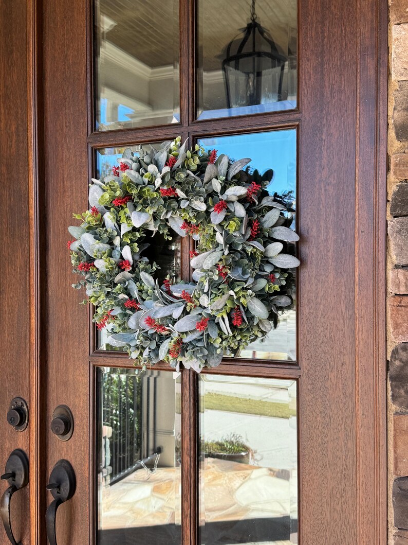 May include: A circular wreath featuring a dense arrangement of soft, sage-green lamb's ear leaves interspersed with small, vibrant red berry clusters and sprigs of green foliage, mounted on a dark brown wooden door with glass panels.