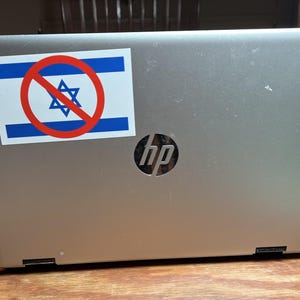 May include: Silver HP laptop with a sticker featuring the Israeli flag crossed out with a red circle and slash. The laptop is on a wooden table.