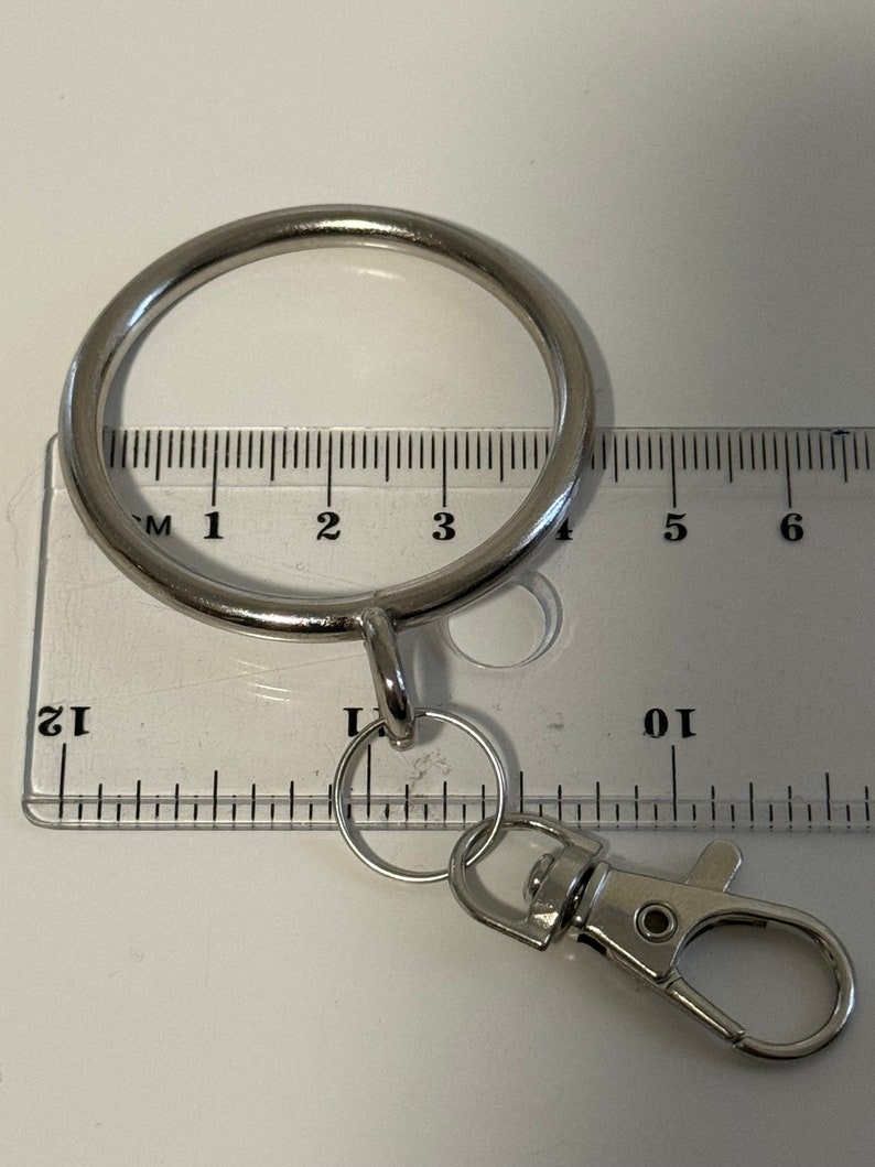 Puede incluir: Anillo de llaves plateado con un anillo redondo grande y un anillo m&aacute;s peque&ntilde;o con un clip. El anillo se mide con una regla, mostrando que tiene aproximadamente 10 cm de di&aacute;metro.