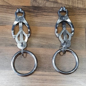 May include: Two silver metal clamps with rings attached. The clamps have a unique design with a small opening at the top and a larger opening at the bottom. The rings are attached to the clamps with a small metal loop.