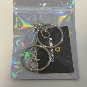Puede incluir: Dos llaveros plateados con un cierre giratorio plateado. Los llaveros est&aacute;n empaquetados en una bolsa de pl&aacute;stico transparente con una tarjeta negra que dice "Kink And Co" en letras doradas.