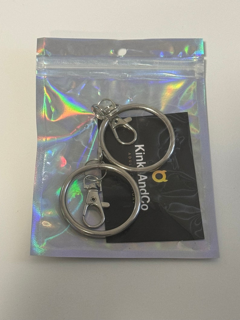Puede incluir: Dos llaveros plateados con un cierre giratorio plateado. Los llaveros est&aacute;n empaquetados en una bolsa de pl&aacute;stico transparente con una tarjeta negra que dice "Kink And Co" en letras doradas.