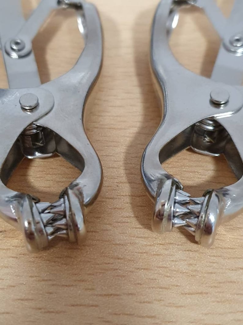 May include: Two silver metal clamps with a textured, twisted design on the ends. The clamps are closed and are lying on a light brown wooden surface.
