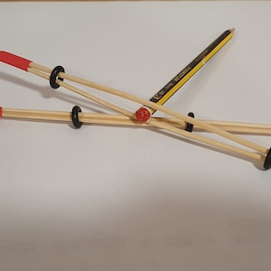 May include: Two wooden sticks with red tips are connected by black rubber bands and a yellow pencil. The pencil is held in place by the rubber bands and sticks.