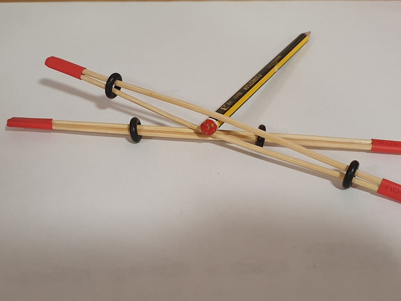 May include: Two wooden sticks with red tips are connected by black rubber bands and a yellow pencil. The pencil is held in place by the rubber bands and sticks.