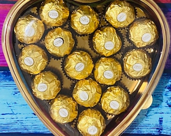 May include: A heart-shaped box filled with golden chocolate candies. The box is made of clear plastic and has a gold trim. The candies are wrapped in gold foil and have the words "Ferrero Rocher" printed on them.