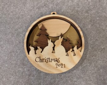 Peut inclure: Une décoration de Noël en bois avec un design en couches représentant une lune, des arbres et des lapins. L'ornement est inscrit avec "Christmas 2021".