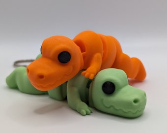 Peut inclure: Un porte-clés en forme de dinosaure vert et orange avec de grands yeux. Le dinosaure orange est sur le dinosaure vert.