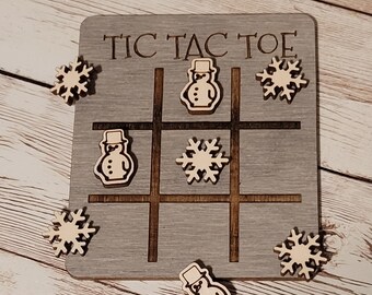 Peut inclure: Un jeu de tic-tac-toe en bois avec un fond gris et les mots "Tic Tac Toe" imprimés dessus. Les pièces du jeu sont en bois et présentent un motif de bonhomme de neige et de flocon de neige.