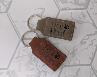 Peut inclure: Deux porte-clés en cuir marron avec des anneaux de clés argentés. Un porte-clés dit "Dog Dad" avec une empreinte de patte. L'autre porte-clés dit "Dog Mom" avec une empreinte de patte.