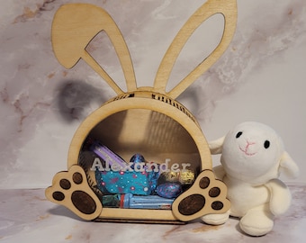 Peut inclure: Un panier de Pâques en forme de lapin en bois avec une fenêtre en plastique transparent. Le panier est rempli d'œufs en chocolat et de bonbons. Le lapin a une empreinte de patte de chaque côté et le nom "Alexander" est écrit sur le devant.