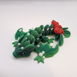 Op de afbeelding: Een groene en rode kerst draak gemaakt van polymer clay. De draak is versierd met witte hulst bessen en heeft een rode strik op zijn staart.