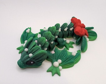 Peut inclure: Un dragon de Noël vert et rouge en pâte polymère. Le dragon est décoré de baies de houx blanches et a un nœud rouge sur sa queue.