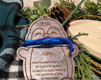 Peut inclure: Une décoration de Noël en bois en forme de pingouin avec une écharpe bleue et grise. La décoration est personnalisée avec le nom "Sophia" et l'année "2022". La décoration a un message qui dit : "Accroche ce pingouin à ton arbre, l'écharpe est aussi grande que moi. Alors sors-le chaque Noël et tu verras que je ne suis plus aussi petit que je l'étais !"