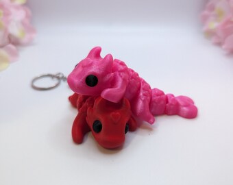 Peut inclure: Un porte-clés en forme de dragon rose et rouge avec un anneau de clé argenté. Les dragons sont petits et ont des yeux noirs. Le dragon rose est sur le dragon rouge.