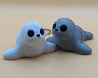 Peut inclure: Deux petites figurines de phoques, l'une blanche et l'autre grise, avec des yeux noirs. Les phoques sont couchés sur une surface brun clair.