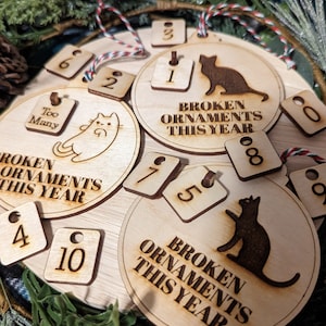 May include: A set of wooden ornaments with a cat theme. The ornaments are round and have the text "Broken Ornaments This Year" on them. There are also smaller square ornaments with numbers on them. The ornaments are decorated with a cat silhouette and the text "Too Many".