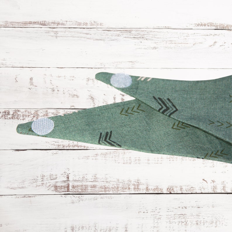May include: A green bandana with a geometric pattern of arrows. The bandana has two white adhesive dots on the corners.