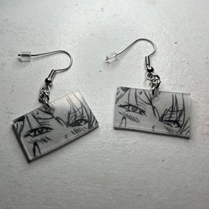 May include: A pair of silver earrings with clear rectangular charms featuring black and white anime-style drawings of eyes.