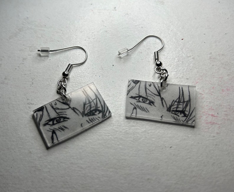 May include: A pair of silver earrings with clear rectangular charms featuring black and white anime-style drawings of eyes.