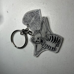 May include: A clear plastic keychain with a black and white skeleton design. The skeleton is holding its ribs in its hands and has a skull with a surprised expression. The keychain has a silver metal ring.