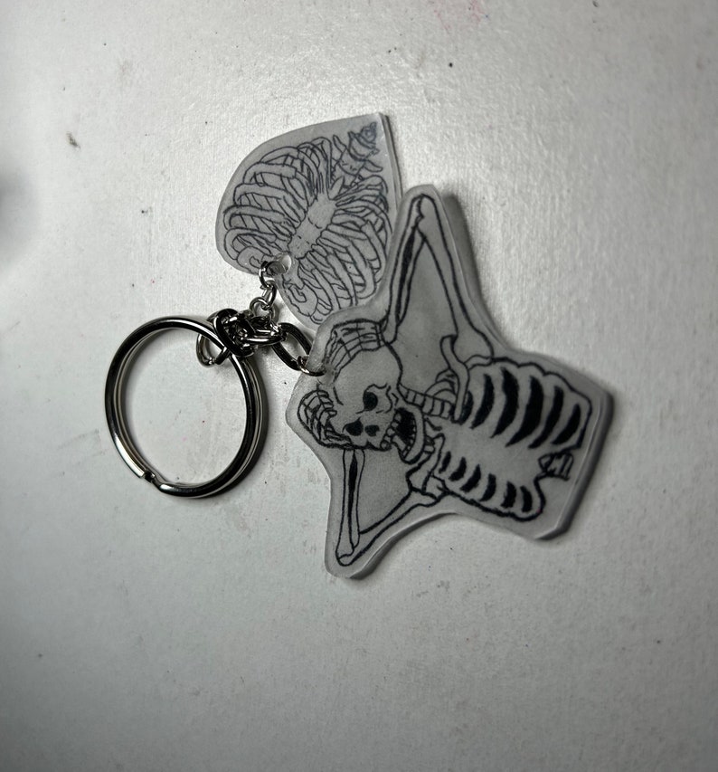 May include: A clear plastic keychain with a black and white skeleton design. The skeleton is holding its ribs in its hands and has a skull with a surprised expression. The keychain has a silver metal ring.