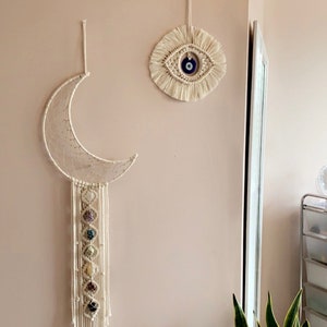 Op de afbeelding: Twee macramé wandhangers, een in de vorm van een halve maan met kristallen die eraan hangen, en de andere een cirkel met een blauw boze oog in het midden.