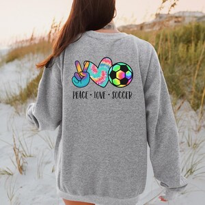May include: Gray sweatshirt with a tie-dye peace sign, heart, and soccer ball graphic on the back. The text "Peace Love Soccer" is printed below the graphic.