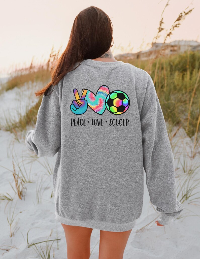 May include: Gray sweatshirt with a tie-dye peace sign, heart, and soccer ball graphic on the back. The text "Peace Love Soccer" is printed below the graphic.