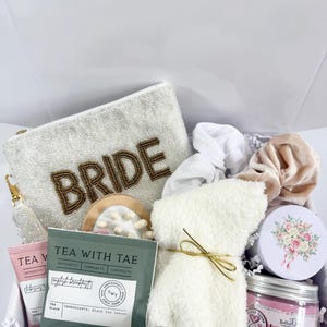 Op de afbeelding: Een bruidsgeschenkdoos met een witte, met kralen bezette clutch met "BRIDE" in gouden kralen, theezakjes, lippenbalsem, een witte pluche badjas, een pot body butter en een bloemig versierde blikken doos.