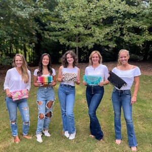 May include: Five women standing in a grassy area, each holding a different beaded clutch bag. The bags are in various colors and patterns, including rainbow, leopard print, turquoise, and black.