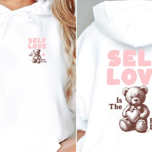 K&ouml;nnte beinhalten: Wei&szlig;er Hoodie mit einem pinkfarbenen "Self Love is the Best Love"-Motiv auf der Vorderseite und einem pinkfarbenen "Self Love"-Motiv auf der R&uuml;ckseite. Das Motiv zeigt einen braunen Teddyb&auml;r, der ein Herz h&auml;lt.