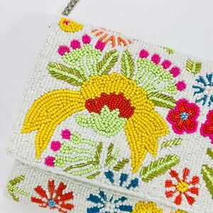 Pu&ograve; includere: Una pochette bianca in perline con un motivo floreale. I fiori sono gialli, arancioni, rossi, rosa, verdi e blu. La borsa ha una tracolla a catena argentata.