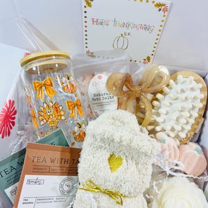 Könnte beinhalten: Eine Thanksgiving-Geschenkbox gefüllt mit Artikeln wie einem Glasbecher mit Strohhalm, einer Tüte Tee, einem flauschigen weißen Hausschuh, Badesalz, einer Badebombe, einer weißen Kerze und einer Holzbürste. Die Box ist mit einer Karte verziert, auf der "Happy Thanksgiving" steht.