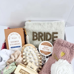 Op de afbeelding: Een bruidsgeschenkdoos met een witte, met kralen bezette clutch met "BRIDE" in gouden kralen, douche-stomers, body butter, lippenbalsem, een haarklem, scrunchies, een badspons en zachte sokken.