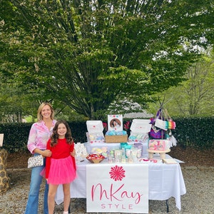 May include: Two people stand in front of a table with a white tablecloth. The table is decorated with various items, including candles, soaps, and other handmade goods. A banner with the text "mKay STYLE" is displayed on the front of the table.