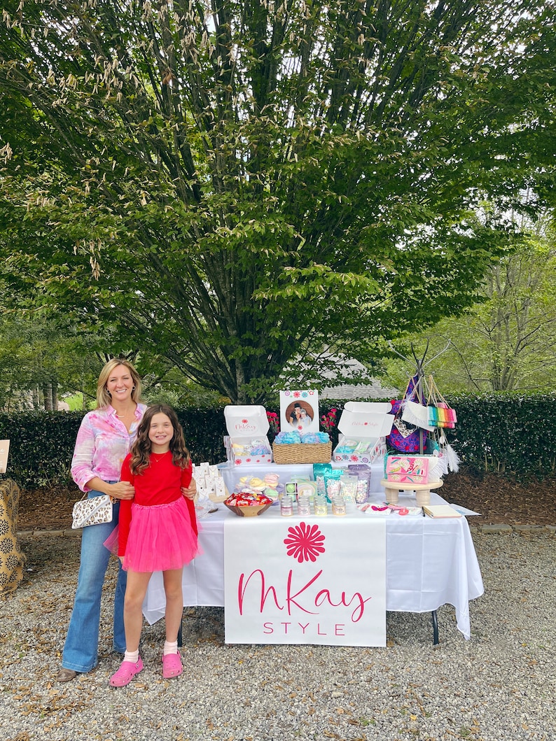 May include: Two people stand in front of a table with a white tablecloth. The table is decorated with various items, including candles, soaps, and other handmade goods. A banner with the text "mKay STYLE" is displayed on the front of the table.