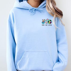 May include: A light blue hoodie with a pocket on the front. The hoodie has a graphic of a peace sign, a heart, and a soccer ball with the text "Peace - Love - Soccer".