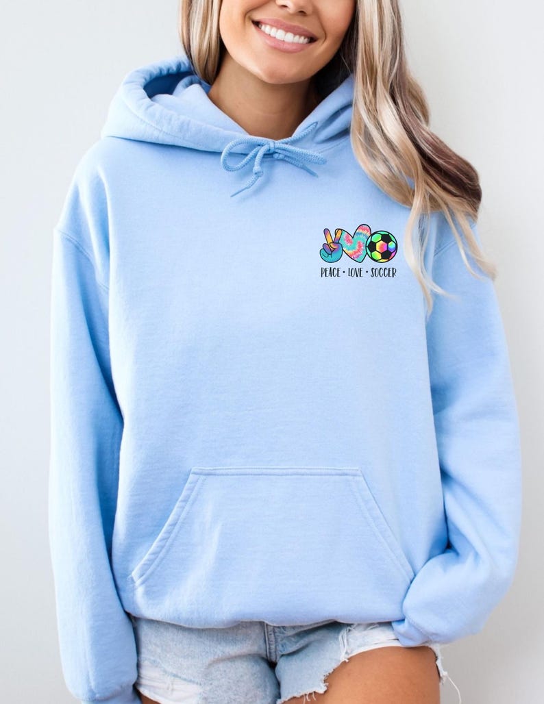 May include: A light blue hoodie with a pocket on the front. The hoodie has a graphic of a peace sign, a heart, and a soccer ball with the text "Peace - Love - Soccer".
