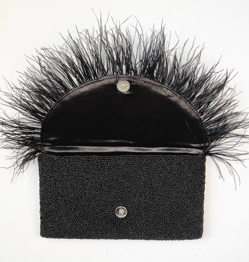 May include: A black beaded clutch with a black feather trim. The clutch has a half-circle flap with a snap closure.